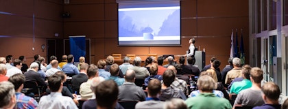 A speaker stands in front of a crowded lecture hall and refers to a presentation projected on a large screen 