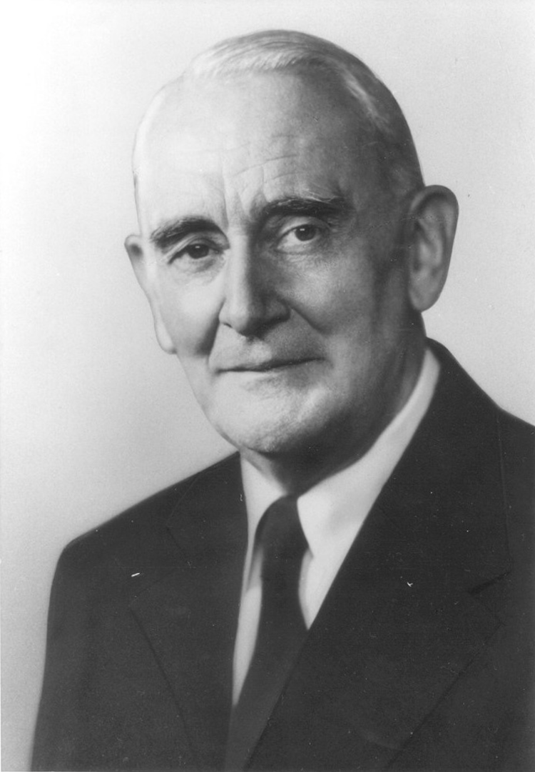 A black and white photo depicts Frank Schofield, who sits against a white backdrop wearing a dark suit.